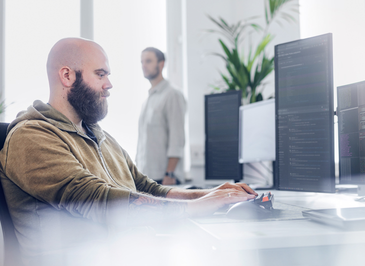 Mann mit Bart arbeitet konzentriert am Computer mit mehreren Monitoren in einem modernen Büro; im Hintergrund steht eine weitere Person.