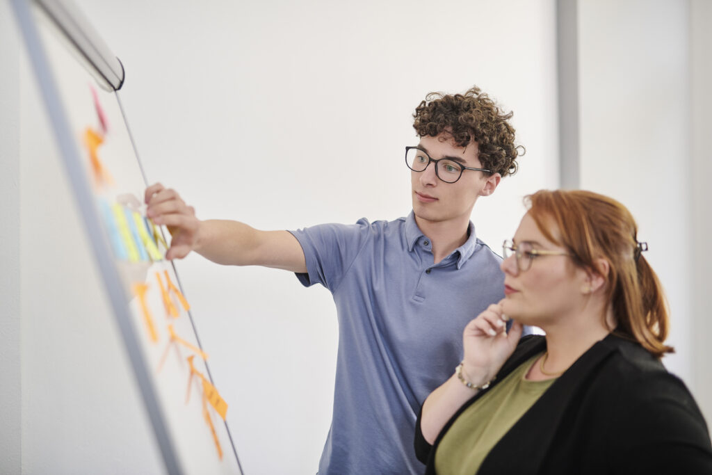 Ein junger Mann zeigt auf bunte Haftnotizen an einem Whiteboard, während eine Frau neben ihm nachdenklich zuschaut.