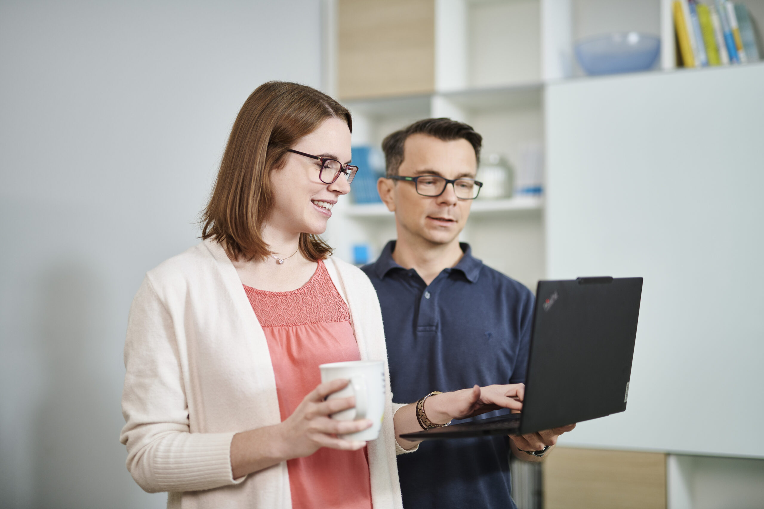 Eine Frau mit Brille hält eine weiße Tasse und lächelt, während sie neben einem Mann mit Brille steht, der auf einen Laptop zeigt. Beide schauen auf den Bildschirm.