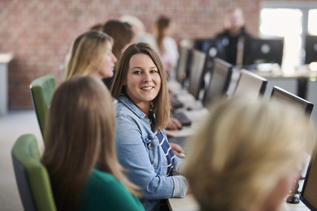 Eine Frau sitzt in einem Computerraum, dreht sich um und lächelt in Richtung Kamera.