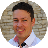 Porträt eines lächelnden Mannes mit dunklen Haaren, weißem Hemd und hellem Hintergrund aus Natursteinen.