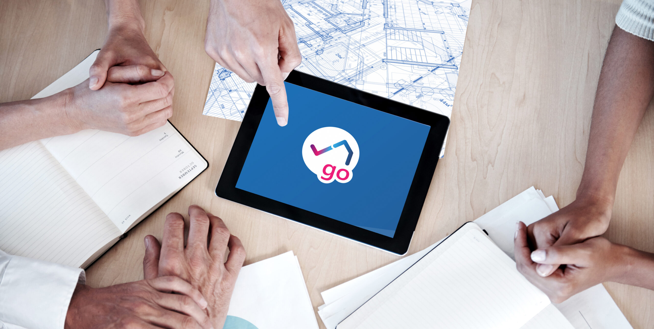 Teammeeting mit Tablet und Bauplänen – Hände zeigen auf Bildschirm mit NeoGo-Logo, Notizbücher und Unterlagen auf Holztisch