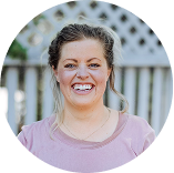 Porträt einer lächelnden Person im rosafarbenen Oberteil vor einem weißen Zaun im Freien.