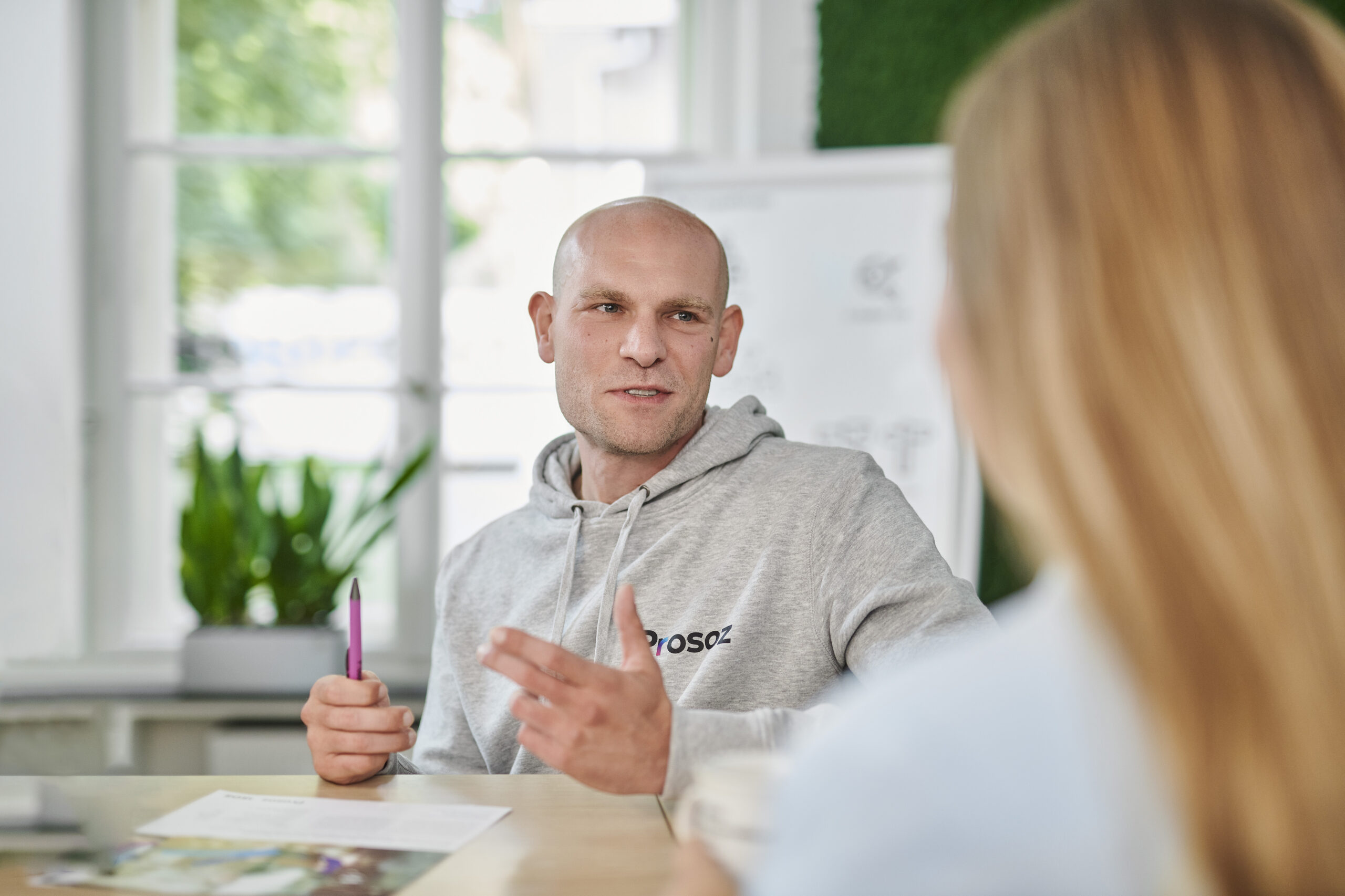 Ein Mann im grauen Hoodie sitzt an einem Tisch, hält einen Stift in der Hand und spricht mit einer weiteren Person