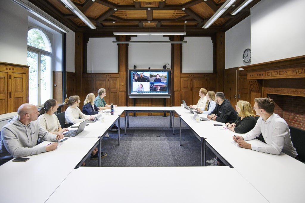 Mehrere Personen sitzen in einem Konferenzraum an zwei langen Tischen und nehmen an einer Videokonferenz teil.