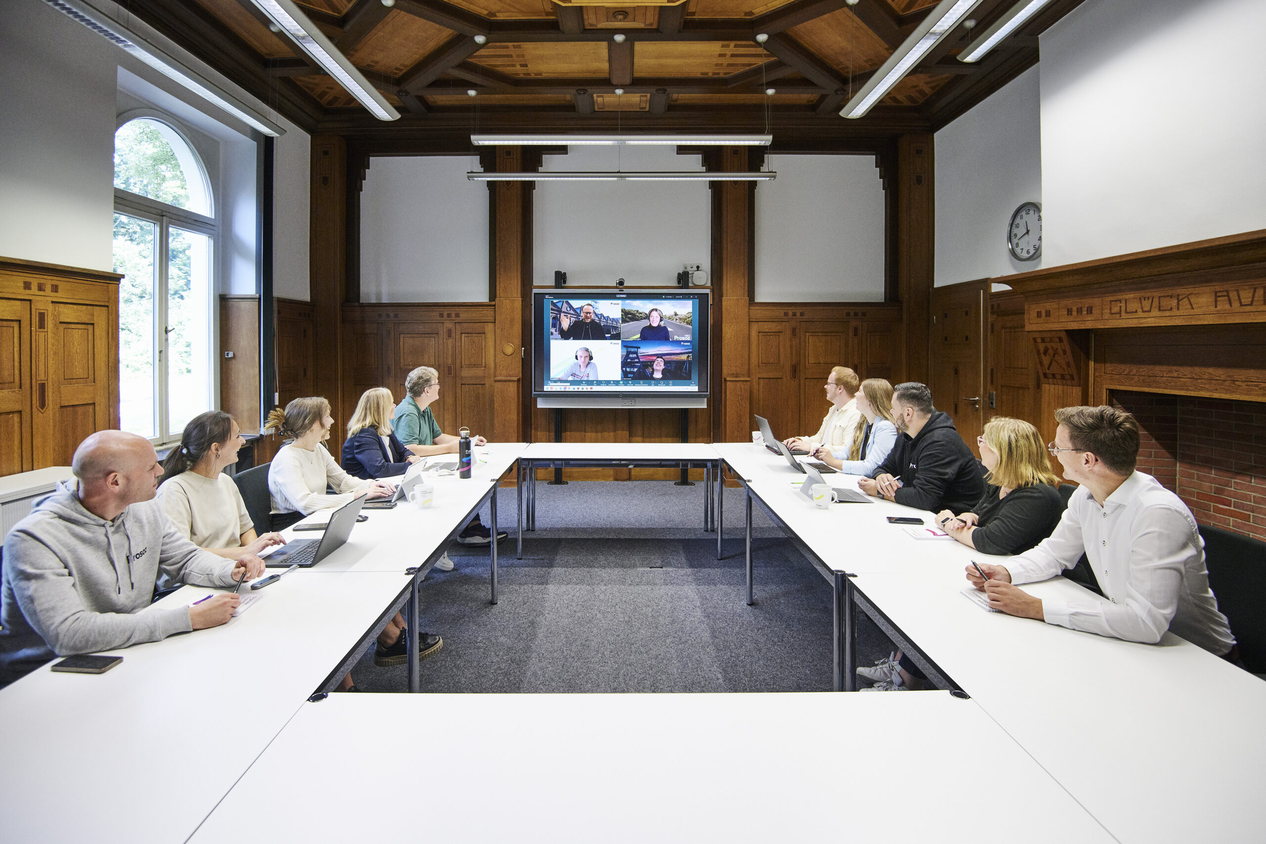 Mehrere Personen sitzen in einem Konferenzraum an zwei langen Tischen und nehmen an einer Videokonferenz teil.
