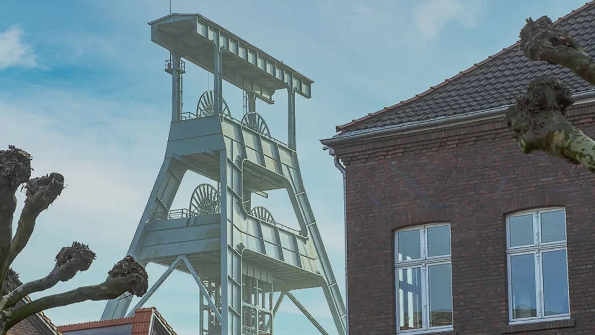 Historischer Förderturm aus Stahl neben einem roten Backsteingebäude unter blauem Himmel.