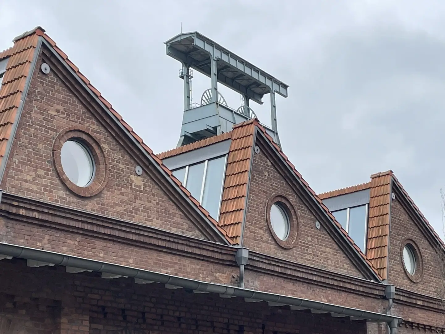 Backsteingebäude mit roten Dachziegeln und runden Fenstern in den Giebeln. Dahinter ragt ein moderner Förderturm aus Stahl in den grauen Himmel.