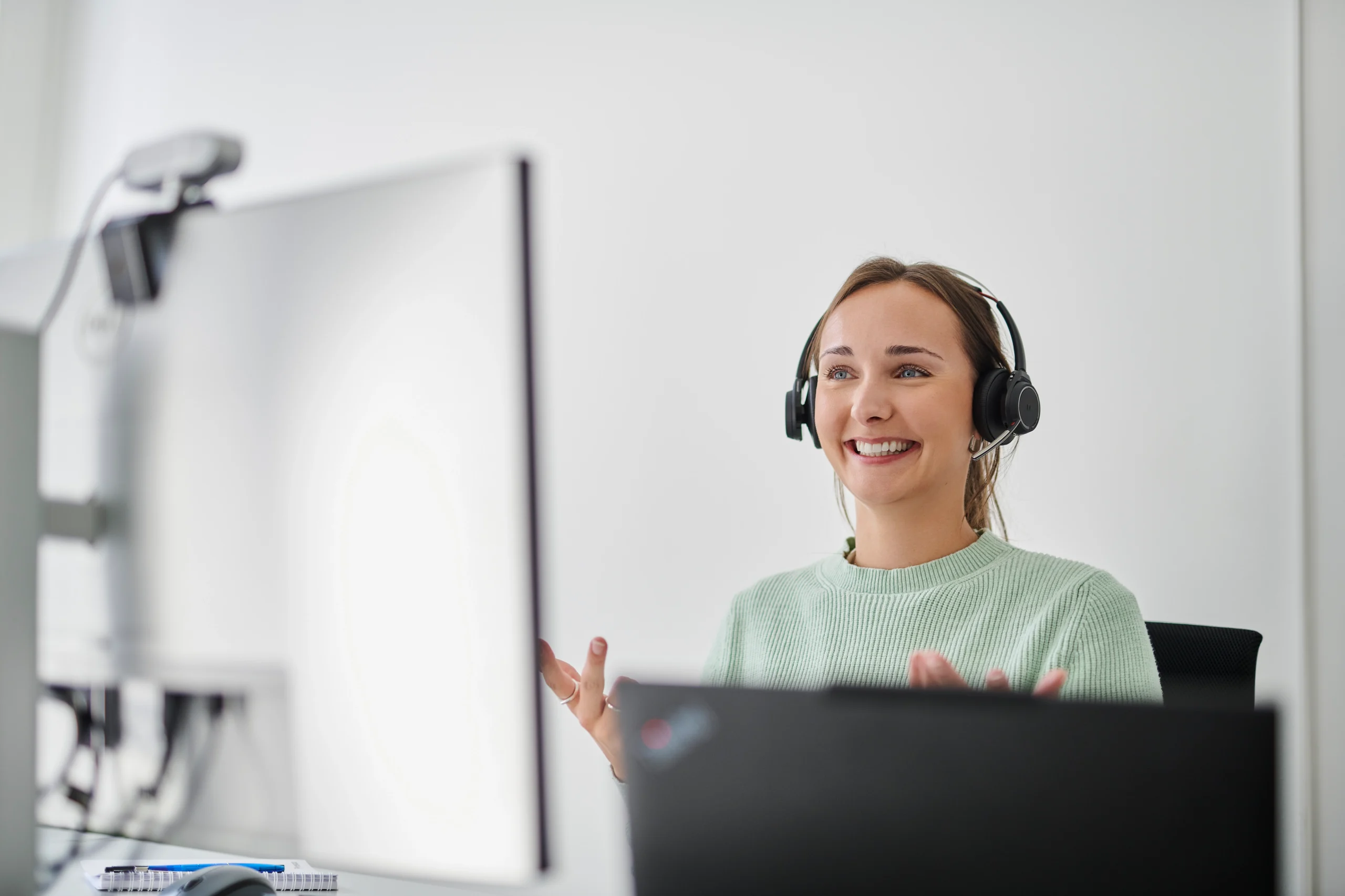 Mitarbeiterin mit Headset spricht lächelnd vor zwei Monitoren und Laptop im Büro.