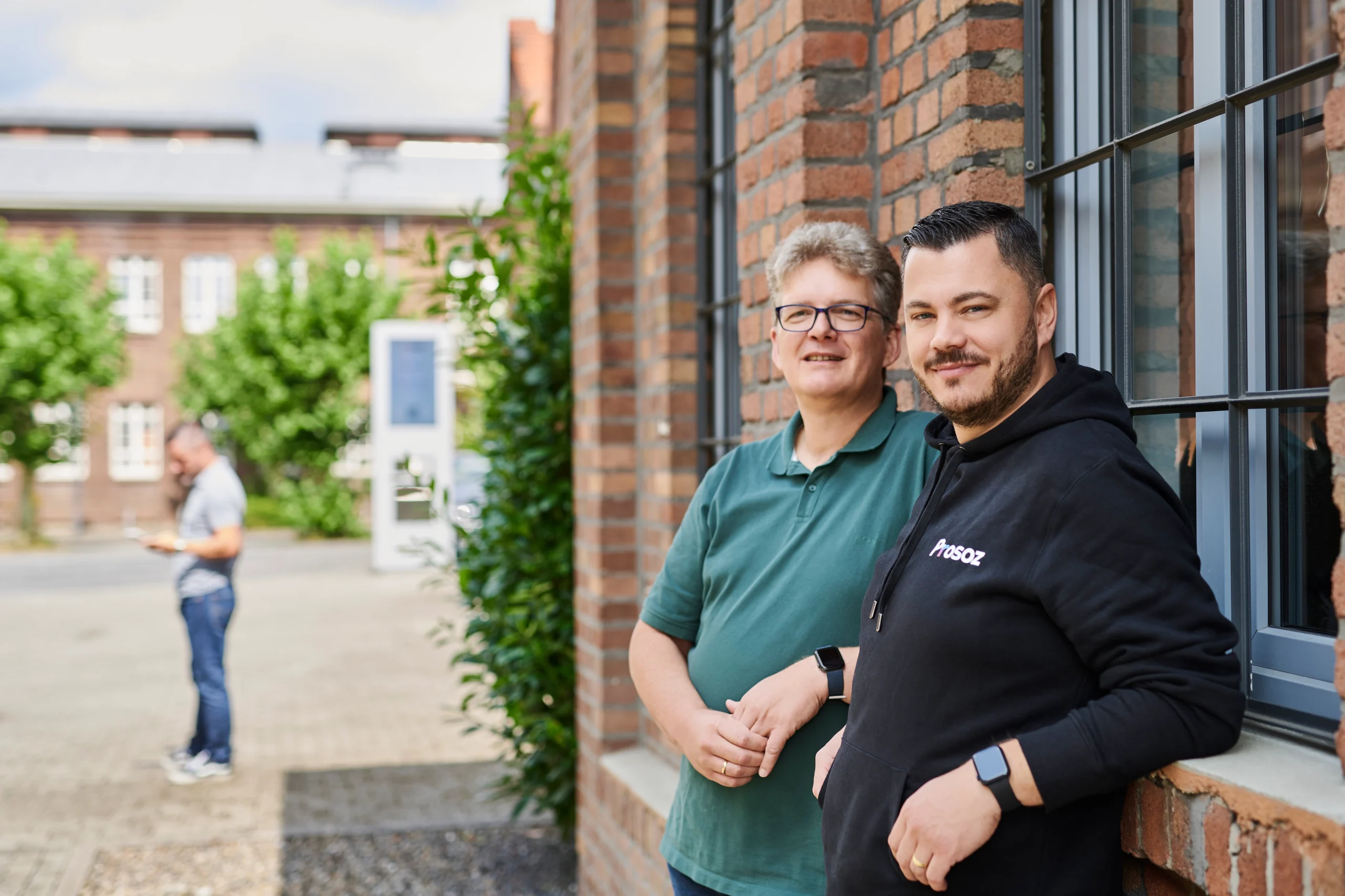 Zwei Kolleg*innen lehnen entspannt an einer Backsteinwand vor Bürofenstern; Campus im Hintergrund.