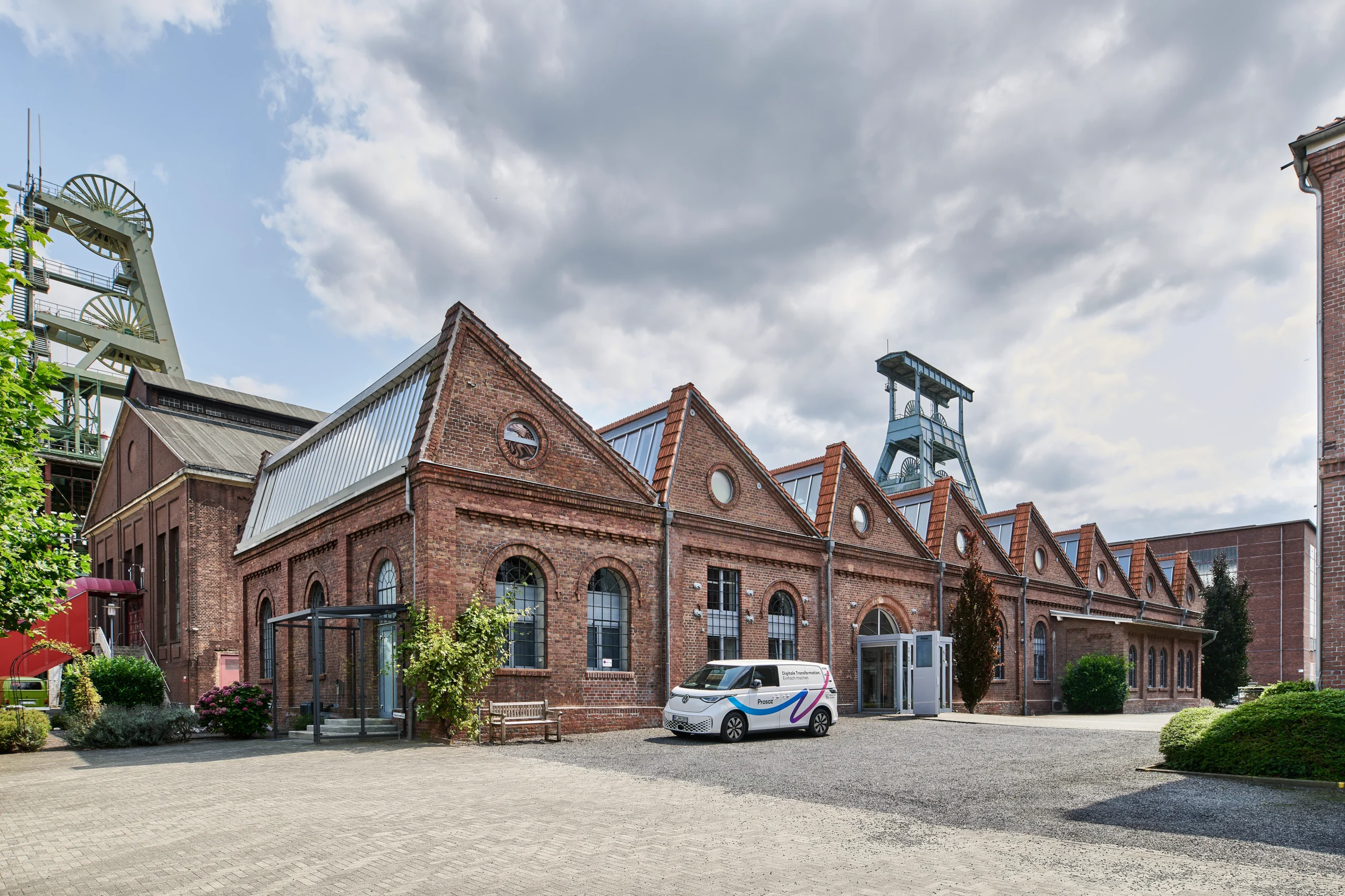 Panorama historischer Backsteingebäude mit Sheddächern; Förderturm und Firmenfahrzeug auf dem Hof.