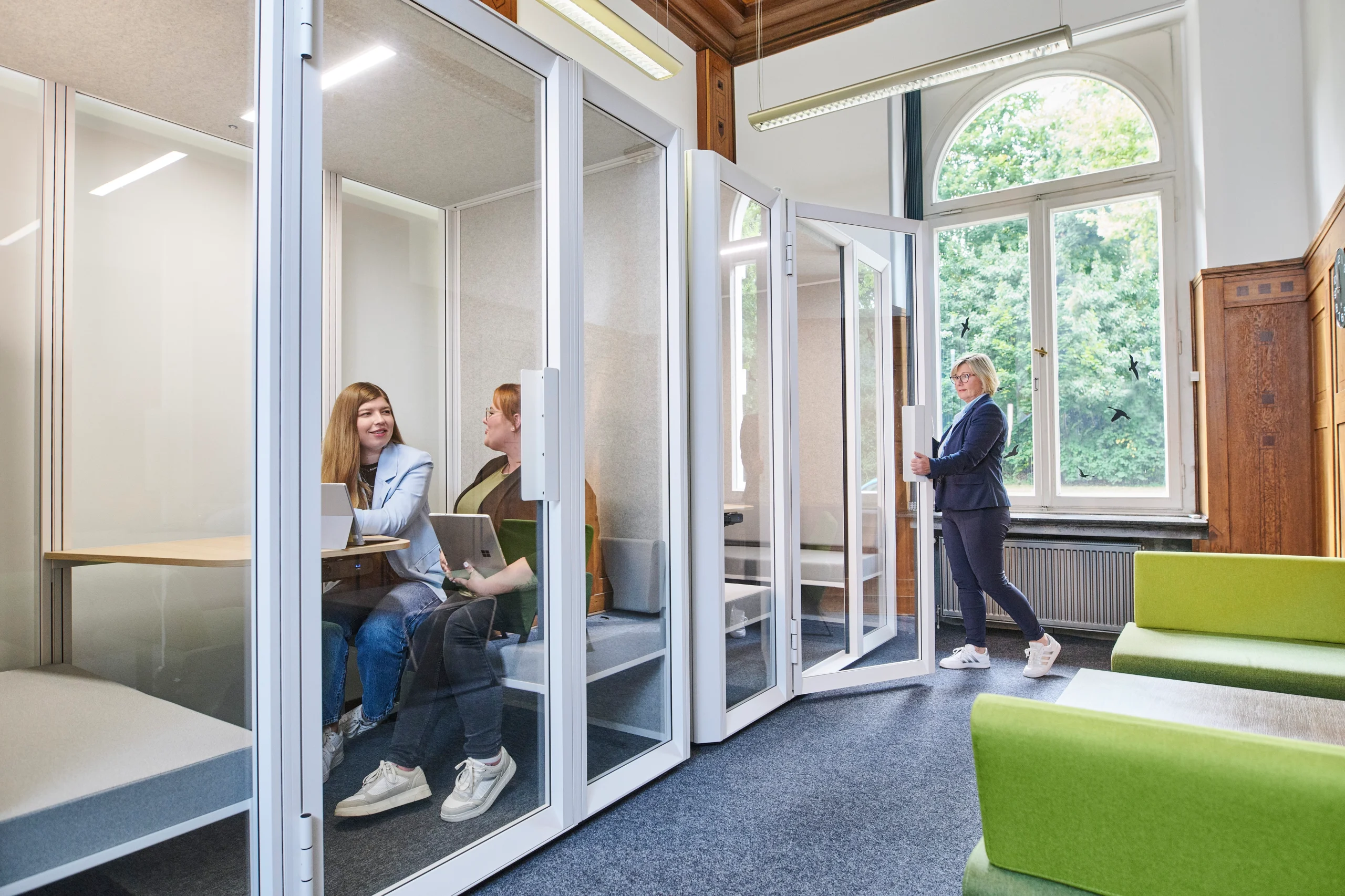 Zwei Kolleginnen arbeiten in schallgedämmten Kabinen; weitere Person öffnet eine Nachbarkabine.