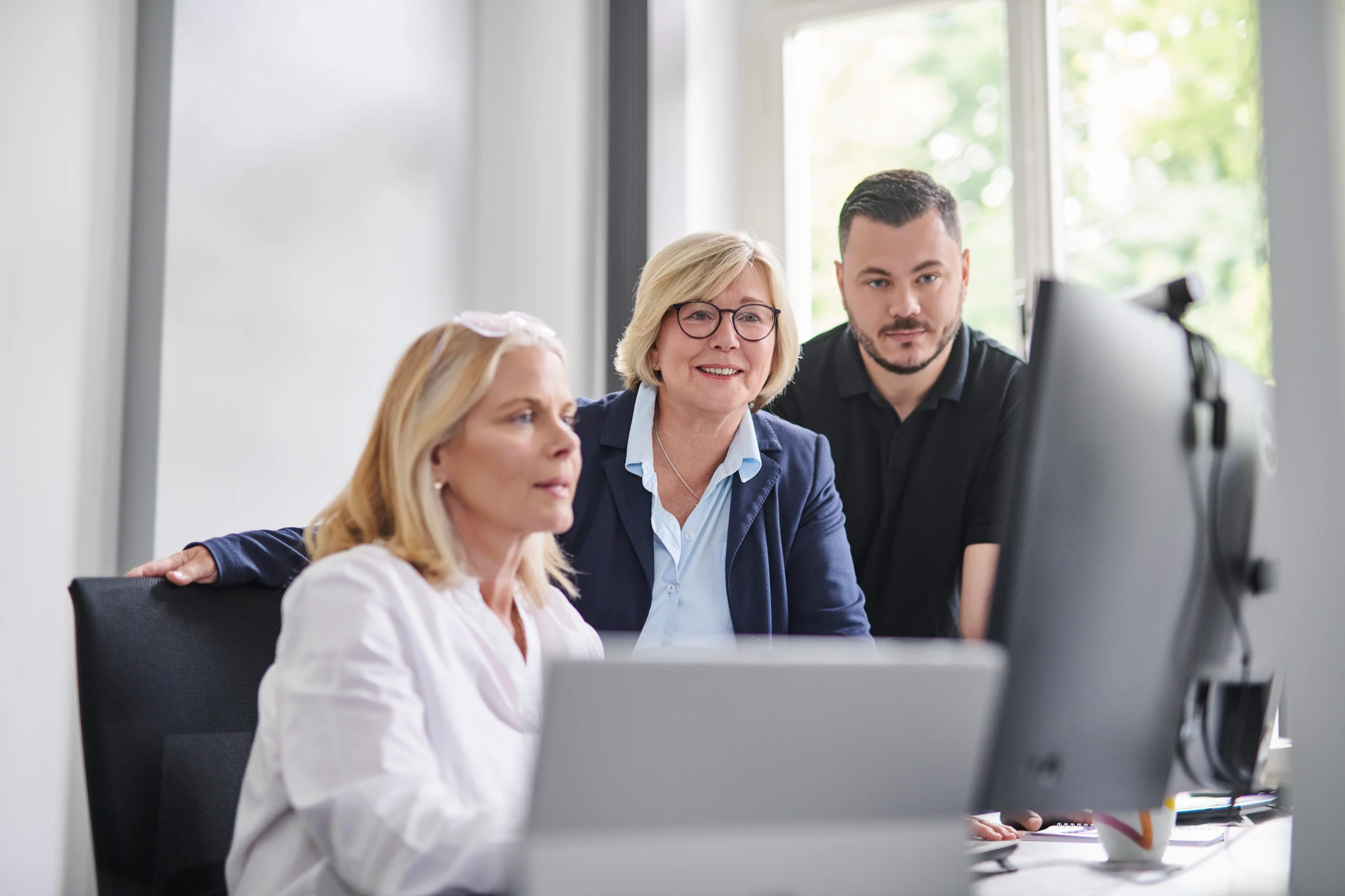 Drei Kolleg*innen schauen konzentriert auf einen großen Monitor; Tageslichtbüro.