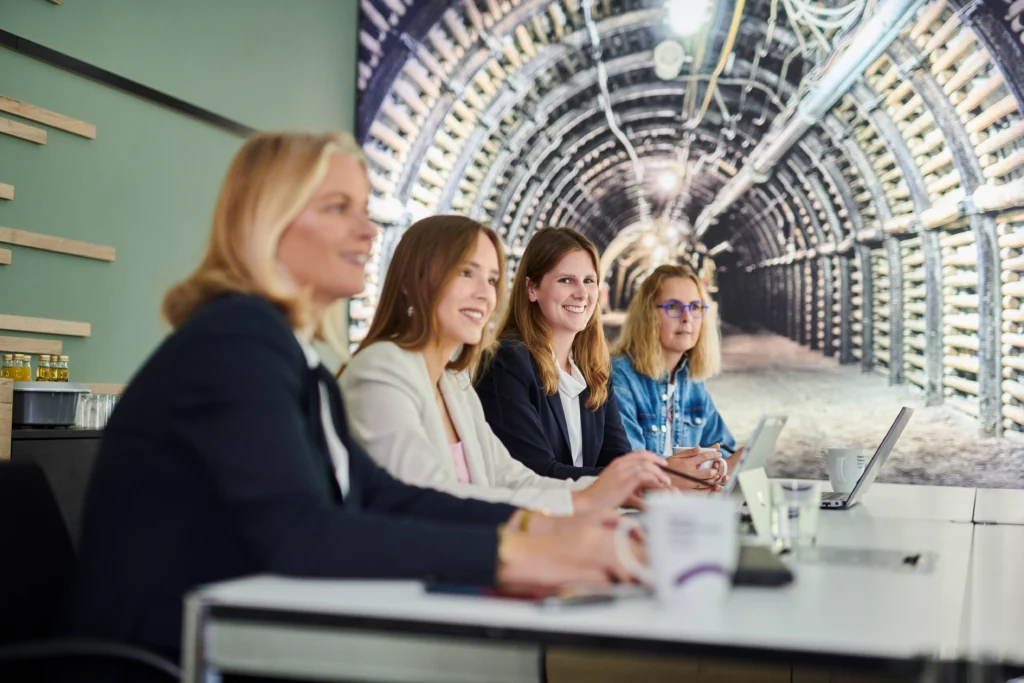 Vier Frauen sitzen lächelnd am Konferenztisch; große Wandgrafik zeigt Stollenröhre.