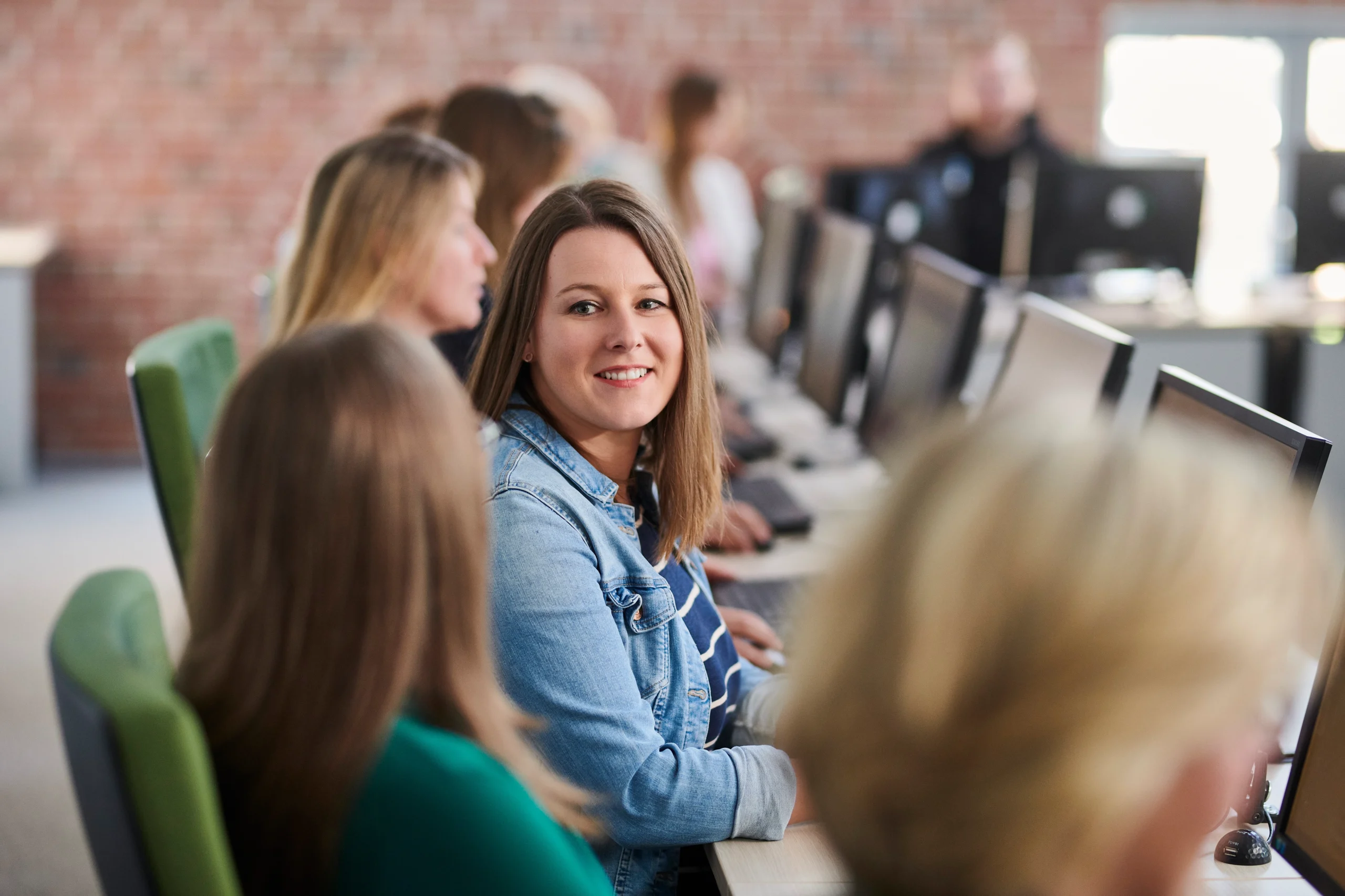 Teilnehmende sitzen an einer Reihe von PCs; Frau in Jeansjacke dreht sich lächelnd um.