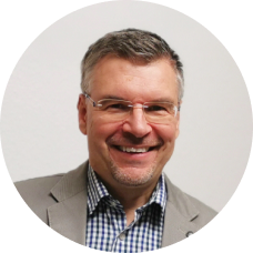 Mann mit Brille und Bart trägt kariertes Hemd und graues Sakko – sympathisches Business-Porträt vor neutralem Hintergrund.